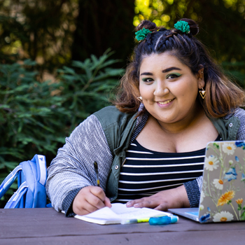smiling student in sweater sitting on wooden bench with her work, laptop and backpack